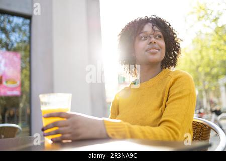 Positive junge afroamerikanische Frau mit lockigem Haar, die lächelt und wegsieht, während sie am Tisch sitzt Stockfoto