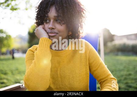 Porträt einer aufmerksamen jungen afroamerikanischen Frau mit der Hand auf die Wange, die wegblickt, während sie auf dem Liegestuhl im grünen Park im Sonnenlicht gegen Unschärfe sitzt Stockfoto
