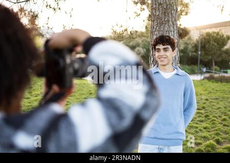 Anonyme Person mit Fotokamera, die ein Bild von einem lächelnden jungen Mann macht, der mit den Händen hinter dem Baum steht und tagsüber in den grünen Park blickt Stockfoto