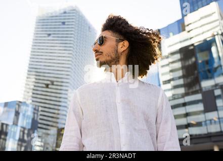 Ein afroamerikanischer Mann mit moderner Kleidung, Afro-Frisur und Sonnenbrille, der auf einer von sonnenbeleuchtetem Bu umgebenen Straße steht Stockfoto