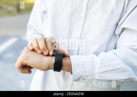 Nicht wiedererkennbarer Mann in weißem Hemd mit Smartwatch, die auf der Straße steht und bei sonnigem Tageslicht die Smartwatch überprüft Stockfoto