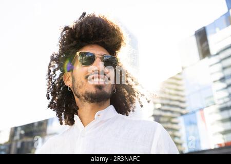 Ein afroamerikanischer Mann mit moderner Kleidung, Afro-Frisur und Sonnenbrille, der auf einem verschwommenen Hintergrund in die Mitte der Stadt blickt Stockfoto