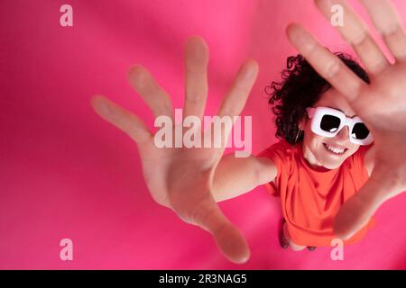 Von oben, von einer fröhlichen jungen Frau in leuchtenden Outfits und einer weißen Sonnenbrille, die Arme mit Handflächen vor der Kamera auf pinkfarbenem Hintergrund hebt Stockfoto