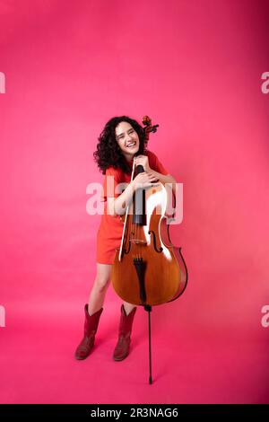 Ein ganzer Körper von aufgeregter junger Musikerin mit lockigem Haar, die ein leuchtendes Kleid mit Stiefeln trägt und im Studio mit pi mit Cello in die Kamera schaut Stockfoto