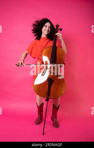 Ein ganzer Körper von aufgeregter junger Musikerin mit lockigem Haar, leuchtendem Kleid, Stiefeln, geschlossenem Blick und Cello-Spielen im Studio w Stockfoto