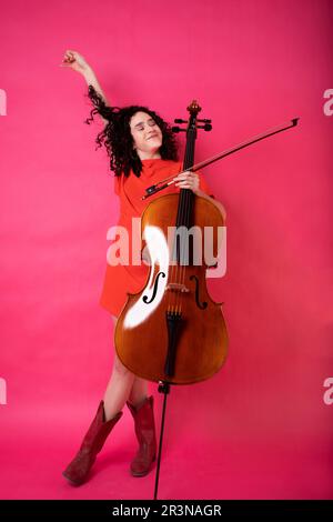 Ein ganzer Körper von aufgeregter junger Musikerin mit lockigem Haar, die ein leuchtendes Kleid mit Stiefeln trägt und mit geschlossenen Augen und mit Cello im Studio steht Stockfoto