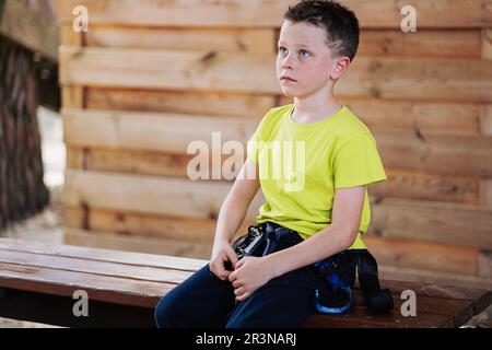 Ein nachdenklicher Schuljunge in heller Kleidung mit Trekkinggürtel, der auf einer Bank an der Holzwand sitzt und beim Warten im Seilpark mit Bedacht wegschaut Stockfoto