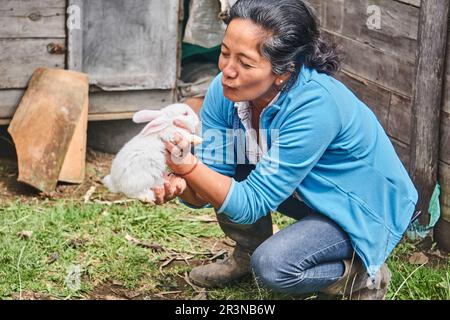 Positiv reife lateinamerikanische Landwirtin mit dunklem Haar in Freizeitkleidung Umarmung und Streichelei niedliches flauschiges kleines Kaninchen, während sie auf dem Fleck sitzt Stockfoto