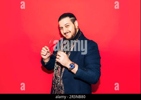 Ein positiver junger Dandy-Typ im trendigen Anzug, mit Seidenschal, Brille in der Hand und Blick in die Kamera, während er im Studio vor rotem Hintergrund steht Stockfoto