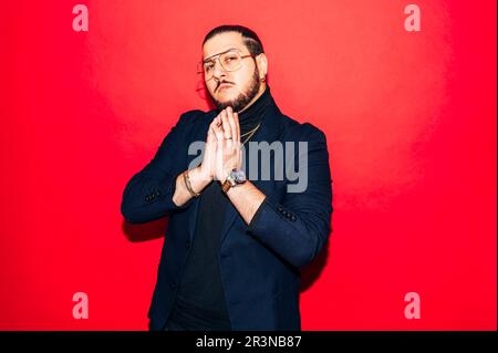 Porträt eines bärtigen Dandy-Mannes in trendigen Outfits und einer Brille mit gefalteten Händen, die auf die Kamera blickt, während sie vor rotem Hintergrund steht Stockfoto