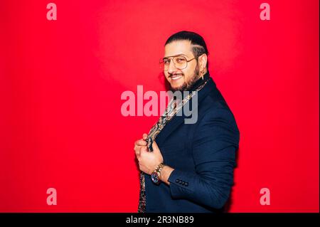 Seitenansicht eines jungen bärtigen Dandys in teurem Outfit mit Seidenschal und Brille, der in die Kamera schaut, während er vor rotem Hintergrund steht Stockfoto