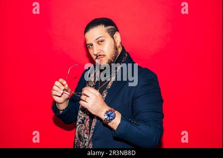 Ein positiver junger Dandy-Typ im trendigen Anzug, mit Seidenschal, Brille in der Hand und Blick in die Kamera, während er im Studio vor rotem Hintergrund steht Stockfoto