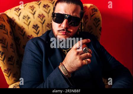 Ein junger Mann mit Bart im Anzug und einer Sonnenbrille sitzt im Sessel mit Zigarre und schaut vor rotem Hintergrund in die Kamera Stockfoto