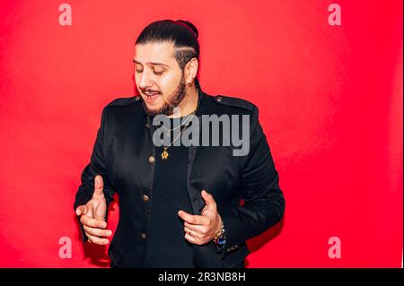 Der junge, bärtige Sänger im schwarzen Anzug mit wegweisenden Accessoires singt ein Lied vor rotem Hintergrund im Studio Stockfoto