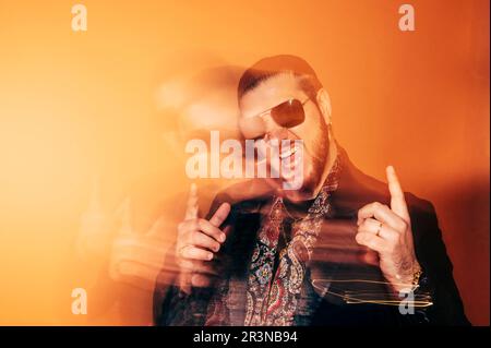 Ein unscharfer bärtiger männlicher Hipster in einer trendigen Jacke und einer Sonnenbrille zeigt nach oben, während er im Studio vor orangefarbenem Hintergrund mit offenem Mund steht Stockfoto