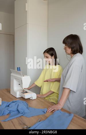Fokussierte Erwachsene Mutter in lässigem T-Shirt, die Teenagern beibringt, mit Nähmaschinen zu arbeiten, während sie neue Kleidung auf einem Holztisch in einem modernen A kreiert Stockfoto