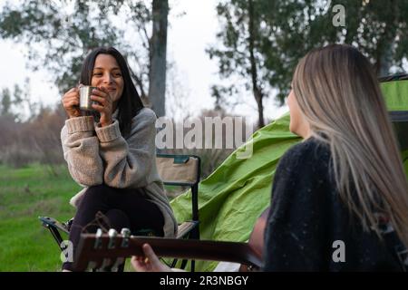 Fröhliche junge Freunde, die lächeln und sich anschauen, sitzen auf Campingstühlen in der Nähe eines Zelts bei Tageslicht und spielen Gitarre im Wald Stockfoto
