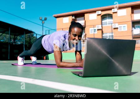 Eine positive, starke Erwachsene, afroamerikanische Sportlerin mit dunklem Haar in Sportbekleidung lächelt, während sie sich Videos über einen Laptop ansieht und Plank-Übungen macht Stockfoto