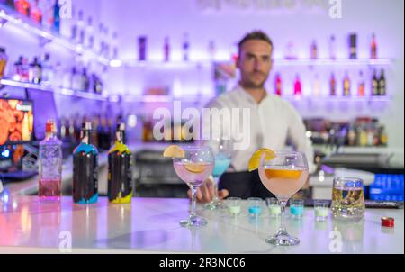Männlicher Barkeeper steht an der Theke mit verschiedenen Cocktailgläsern in verschiedenen Farben zum Servieren und Alkoholflaschen, während er in die Kamera gegen B schaut Stockfoto