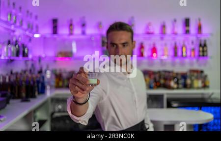 Weicher Fokus des männlichen Barkeepers, der ein Glas süßen Likör anbietet, während er an der Theke in der modernen Bar gegen die verschwommenen lila Regale mit bot steht Stockfoto