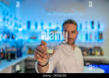 Weicher Fokus des männlichen Barkeepers, der ein Glas süßen Likör anbietet, während er an der Theke in einer modernen Bar gegen verschwommene blaue Regale mit Flasche steht Stockfoto