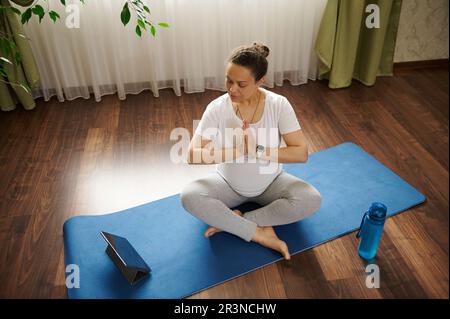 Draufsicht einer schwangeren Frau, die in Lotusstellung sitzt, die Hände zusammenhält und Online-Schwangerschafts-Yoga praktiziert Stockfoto