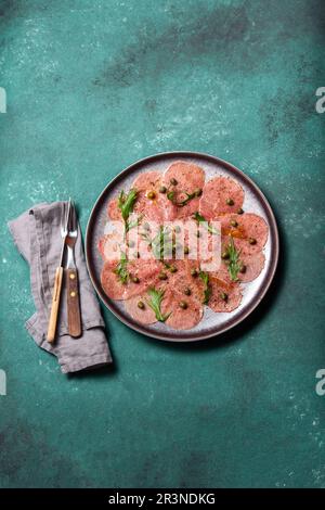 Carpaccio aus Marmorfleisch mit Rucola und Kapern auf grauer Platte, blauer Hintergrund, Draufsicht Stockfoto