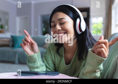 Bild einer glücklichen, birassischen Frau in Kopfhörern, die ihr Smartphone zu Hause auf dem Sofa benutzt, Kopierraum Stockfoto
