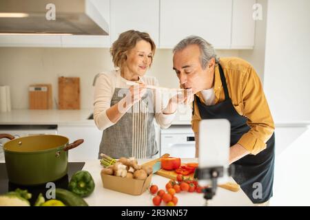 Positive ältere Ehepartner kochen gesundes Abendessen und Frau füttert Ehemann, streamen und verwenden Zellphon in der Küche Stockfoto