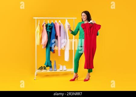 Glückliche Frau, die mit dem Smartphone shoppte und einen Kleiderbügel mit neuem Kleid hielt und Selfie machte, in der Nähe der Kleiderstange posierte Stockfoto