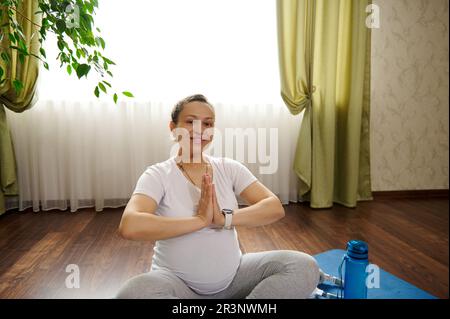 Schwangere Frau lächelt während der Meditation und Yoga-Übung in die Kamera und genießt einen gesunden Lebensstil in der Schwangerschaft. Stockfoto