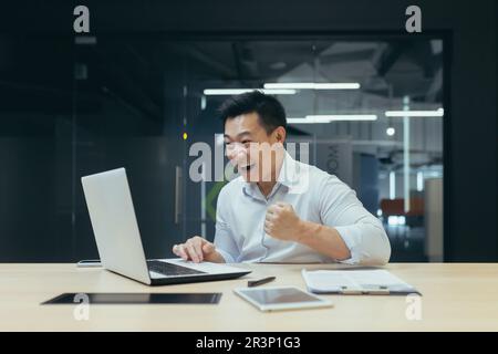 Ein junger asiatischer Geschäftsmann freut sich über einen erfolgreichen finanziellen Deal, ein Projekt. Er arbeitet im Büro am Tisch hinter dem Laptop, schaut auf den Bildschirm und zeigt mit seiner Hand eine Siegergeste. Stockfoto