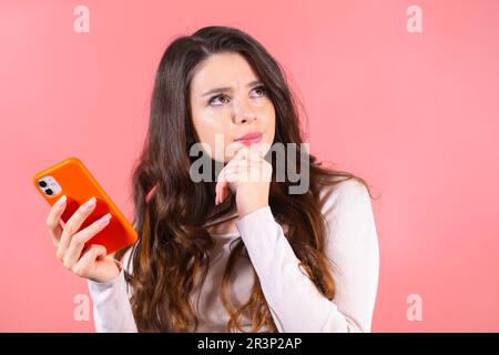 Porträt einer schönen, aufmerksamen Frau mit einem Smartphone auf pinkfarbenem Hintergrund. Schwierige Kaufentscheidung beim Online-Shopping. Stockfoto