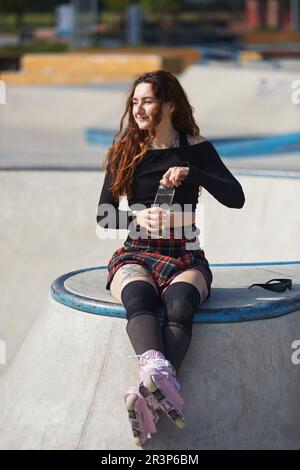 Aggressive Inline-Skaterin mit modernen violetten Rollschuhen, die Wasser aus einer wiederverwendbaren Glasflasche trinkt. Coole tätowierte junge Person, die sich ausruht Stockfoto