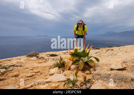 Männlicher Reisender mit Rucksack, der auf einem hohen Berg steht und das Meer überblickt und in die Kamera schaut, Griechenland, Kreta Stockfoto