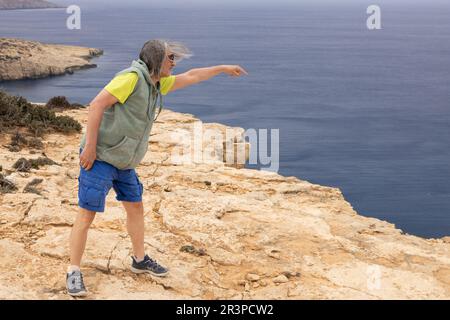 Der grauhaarige Sportler steht auf einer hohen Klippe und zeigt mit seiner Hand auf das Meer, Griechenland, Kreta Stockfoto