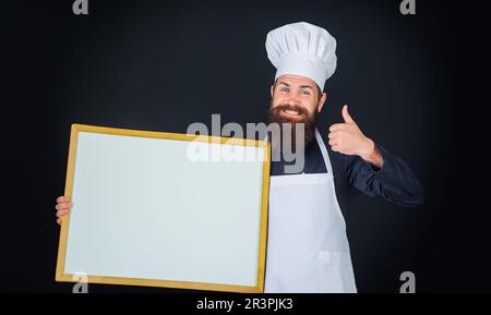 Lächelnder männlicher Koch, Koch oder Bäcker mit leerem Brett mit Daumen nach oben. Koch in Uniform mit leerer Speisekarte an der Tafel. Leeres Plakat für Werbetext Stockfoto