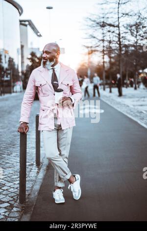 Ein eleganter schwarzer Mann mit kahlem Kopf und einem langen Bart mit Hinweisen auf weißes Haar, trägt eine rosa Jacke und hält fröhlich eine Tasse Kaffee in der Mitte Stockfoto