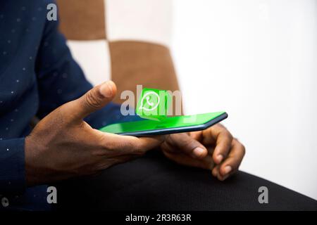 Schwarzes Handheld-Mobiltelefon mit WhatsApp-Logo, das auf dem Bildschirm im Wohnzimmer schwebt. Social-Media-Symbol erscheint aus dem Telefon Stockfoto