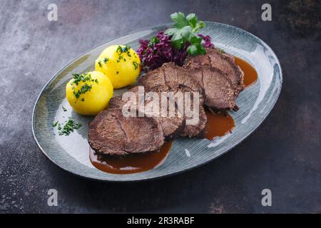 Traditionell geschmorter, marinierter deutscher Sauerbraten aus Rindfleisch mit Blaukraut und Kartoffelknödeln in scharfer dunkler Biersauce serviert Stockfoto