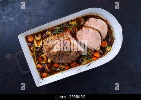 Traditioneller deutscher Schweinebraten, serviert mit Gemüse und Kräutern in dunkler Biersauce, serviert als Draufsicht in einer rustikalen Schmorpfanne Stockfoto