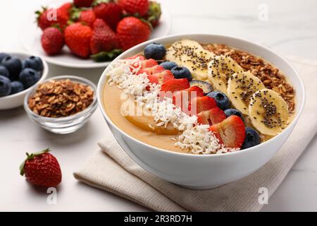 Köstliche Smoothie-Schüssel mit frischen Beeren, Banane, Kokosflocken und Müsli auf weißem Tisch, Nahaufnahme Stockfoto