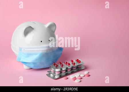 Sparschwein in Gesichtsmaske und Pillen auf rosa Hintergrund, Platz für Text. Krankenversicherung Stockfoto