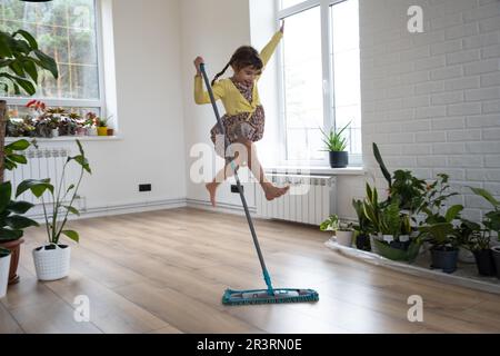 Ein Mädchen tanzt mit einem Mopp, um den Boden in einem neuen Haus zu reinigen - allgemeine Reinigung in einem leeren Raum, die Freude am Umziehen, Hilfe mit dem Stockfoto