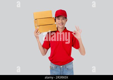 Junger asiatischer Mann in Uniform Rot und Mütze Stehkiste und Geste ok isolierter weißer Hintergrund, Mitarbeiter hält Fracht, Kurier und Zustellung Stockfoto