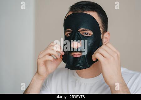 Ein gutaussehender Mann, der eine schwarze Gesichtsmaske auf seinem Gesicht aufträgt und Wellness-Behandlungen erhält. Er pflegt sich selbst. Gesichtsbehandlung, Kosmetologie. Gesichtspflege Stockfoto