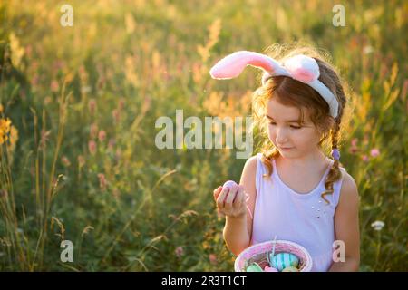 Süßes, lustiges Mädchen mit bemalten Ostereiern im Frühling in der Natur auf einem Feld mit goldenem Sonnenlicht und Blumen. Osterferien, Ostern Stockfoto