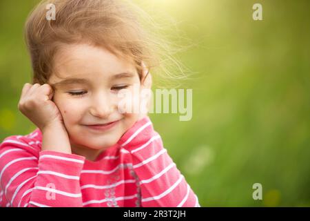 Ein kleines Mädchen in einem korallengestreiften T-Shirt auf grünem Hintergrund auf einem Feld hält ihr Gesicht in den Händen und lächelt sanft. Für Kinder Stockfoto