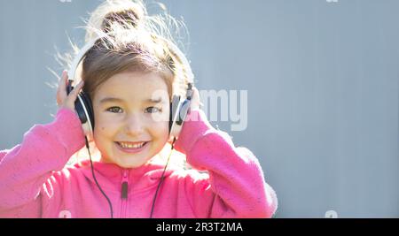 Ein fröhliches Mädchen mit Kopfhörern und einem pinken Hoodie hört Musik und lacht. Draußen, grauer Hintergrund Stockfoto