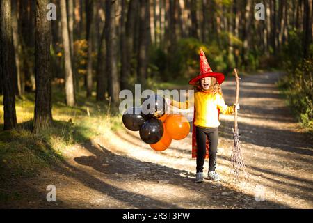 Ein Mädchen in Hexenkostüm und Hut auf einem Besen mit orangefarbenen und schwarzen Ballons spielt im Herbstwald und geht zu einem Hallowee Stockfoto